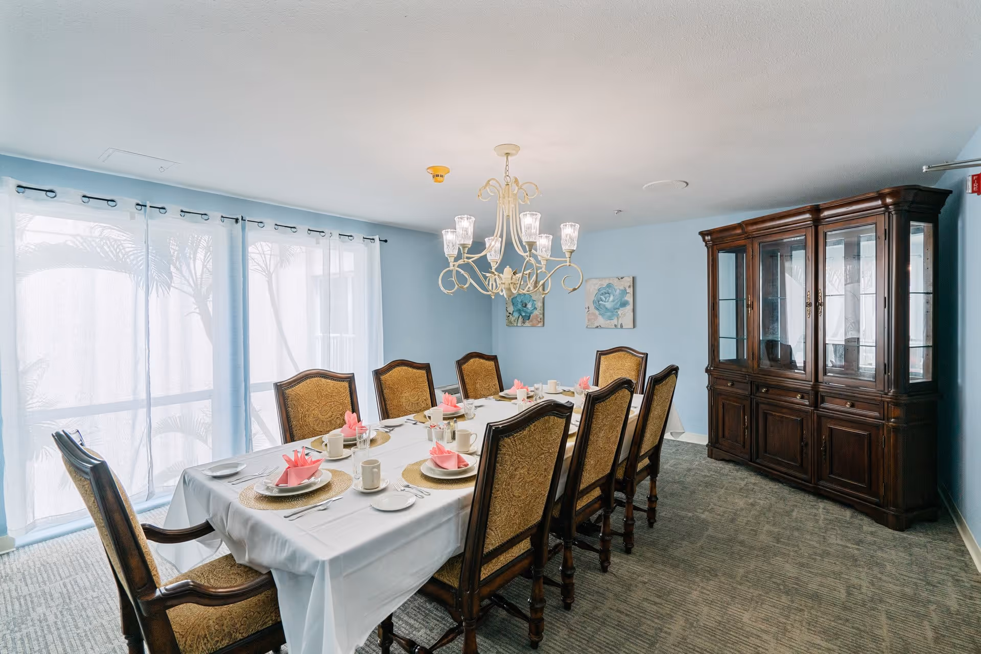 A dining room with a long table set for eight people. The table is covered with a white tablecloth and has plates, cups, and pink folded napkins at each place setting. There are eight upholstered wooden chairs around the table. A chandelier with multiple lights hangs above the table. The room has light blue walls, a large wooden china cabinet, and sheer curtains covering the windows.