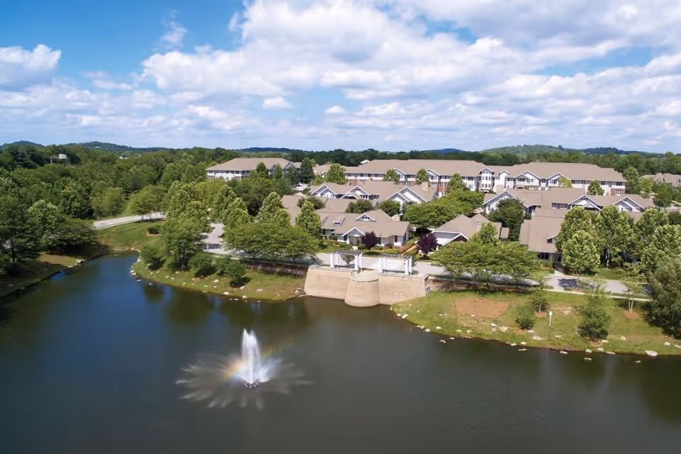 Aerial view of The Heritage at Brentwood senior living facility showing multiple residential buildings with beige roofs surrounded by green trees. In the foreground, there is a pond with a water fountain creating a small rainbow under a partly cloudy sky.