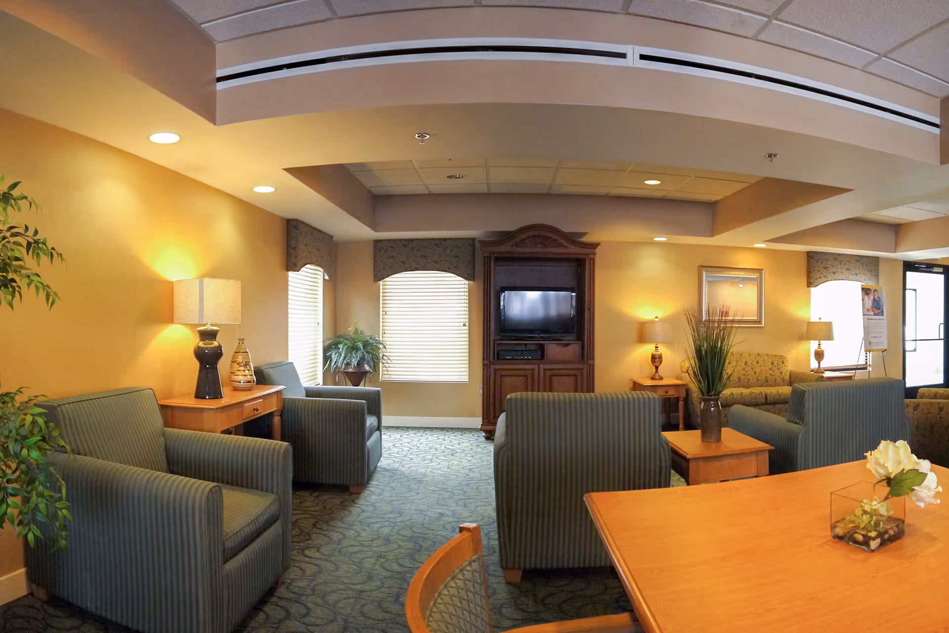A cozy living room area in a senior living facility with several green upholstered armchairs and sofas arranged around wooden side tables and a wooden entertainment center with a TV. The room is warmly lit with table lamps and has large windows with blinds and valances. There are plants and framed artwork on the walls, and a wooden table with a small flower arrangement in the foreground.