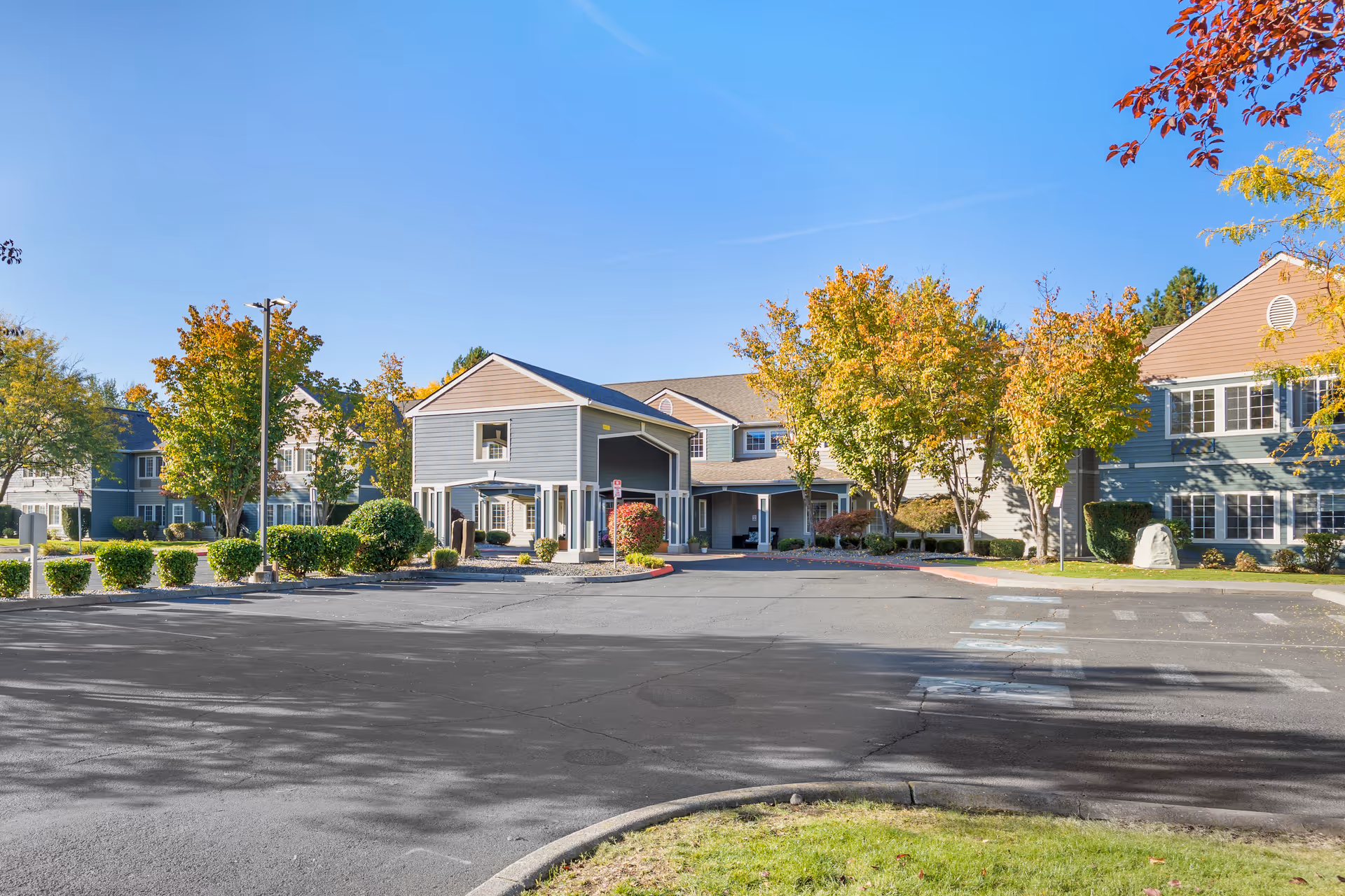 Exterior view of a senior living facility building with a covered entrance, surrounded by trees with autumn foliage and a clear blue sky.