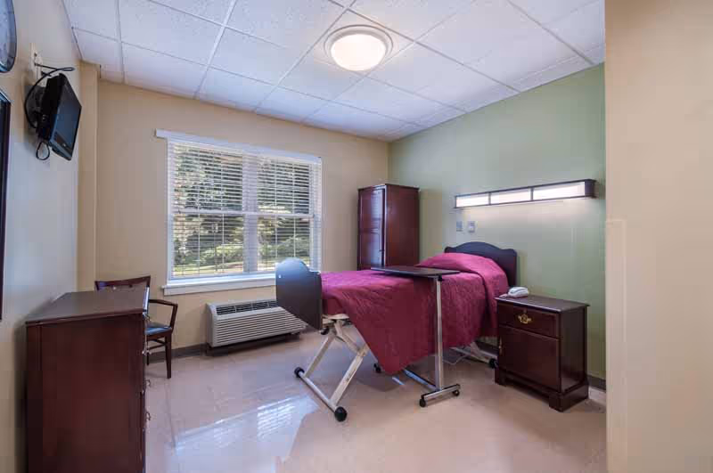 A simple furnished bedroom with an adjustable hospital-style bed covered in a red blanket, nightstand, dresser, and a window overlooking greenery.