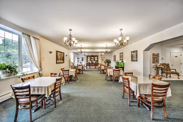 A spacious dining room with multiple wooden tables and chairs arranged neatly, each table covered with a white tablecloth. The room is well-lit with large windows on the left side letting in natural light and several chandeliers hanging from the ceiling. There are framed pictures on the walls, a large wooden cabinet at the far end, and a fish tank on the right side. A cozy seating area with an upholstered chair is visible through an archway on the right.
