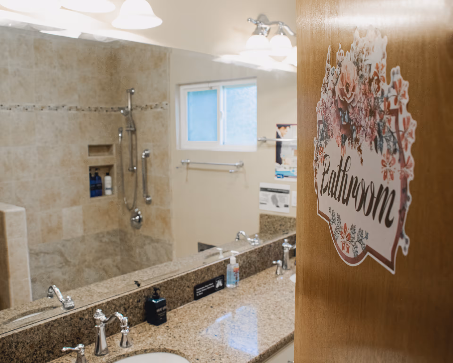 View of a bathroom with a granite countertop featuring two sinks and chrome faucets. A large mirror reflects a tiled shower area with built-in shelves and a handheld showerhead. A wooden door with a floral sign that reads 'Bathroom' is partially open.