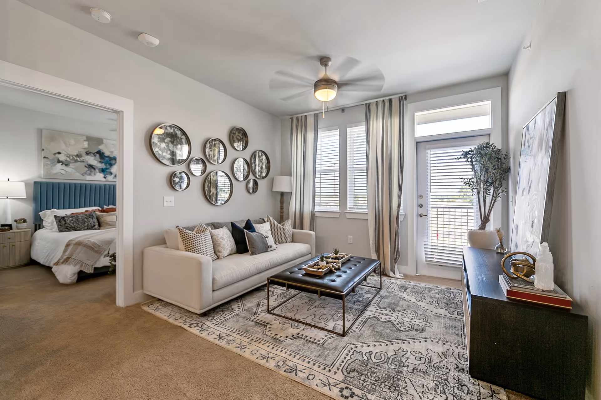 Bright modern living room with a white sofa, multiple round wall mirrors, a tufted ottoman coffee table, patterned rug and a doorway leading to a bedroom.