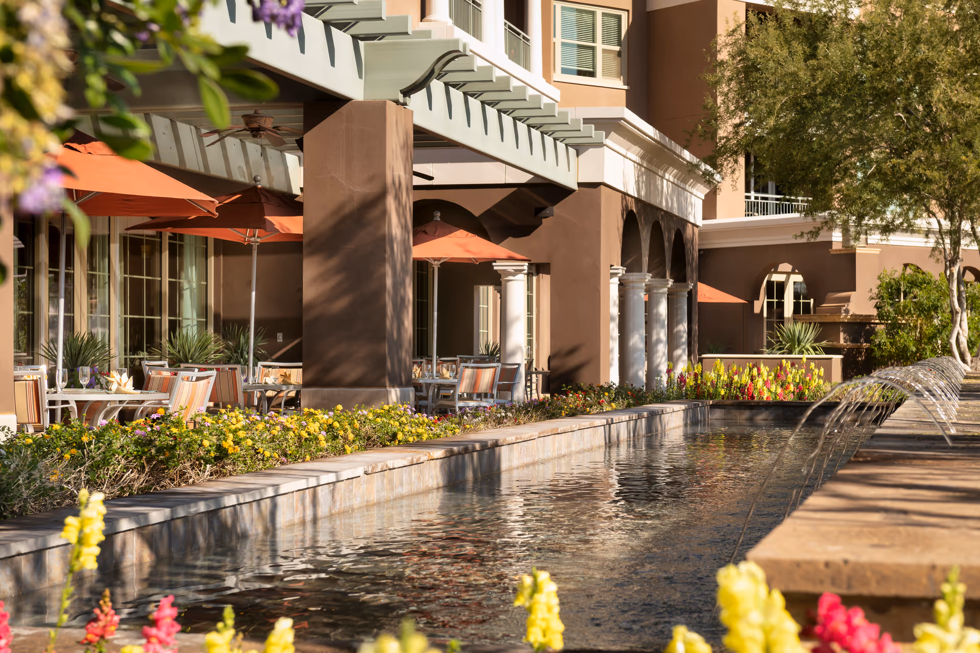 Outdoor courtyard featuring a long reflecting pool with arched water jets, patio seating with orange umbrellas, and flower beds in front of a multi-story building.