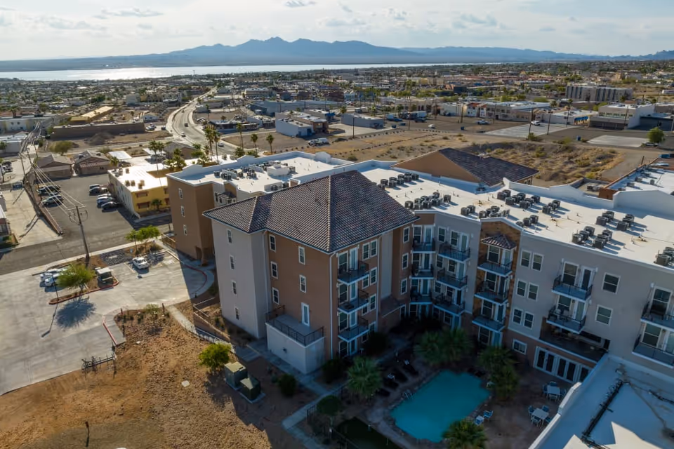 Aerial view of The Views at Lake Havasu Independent Living facility showing a multi-story building with balconies, a swimming pool surrounded by palm trees, and a parking area. The background includes a road, other buildings, and a large body of water with mountains in the distance under a partly cloudy sky.