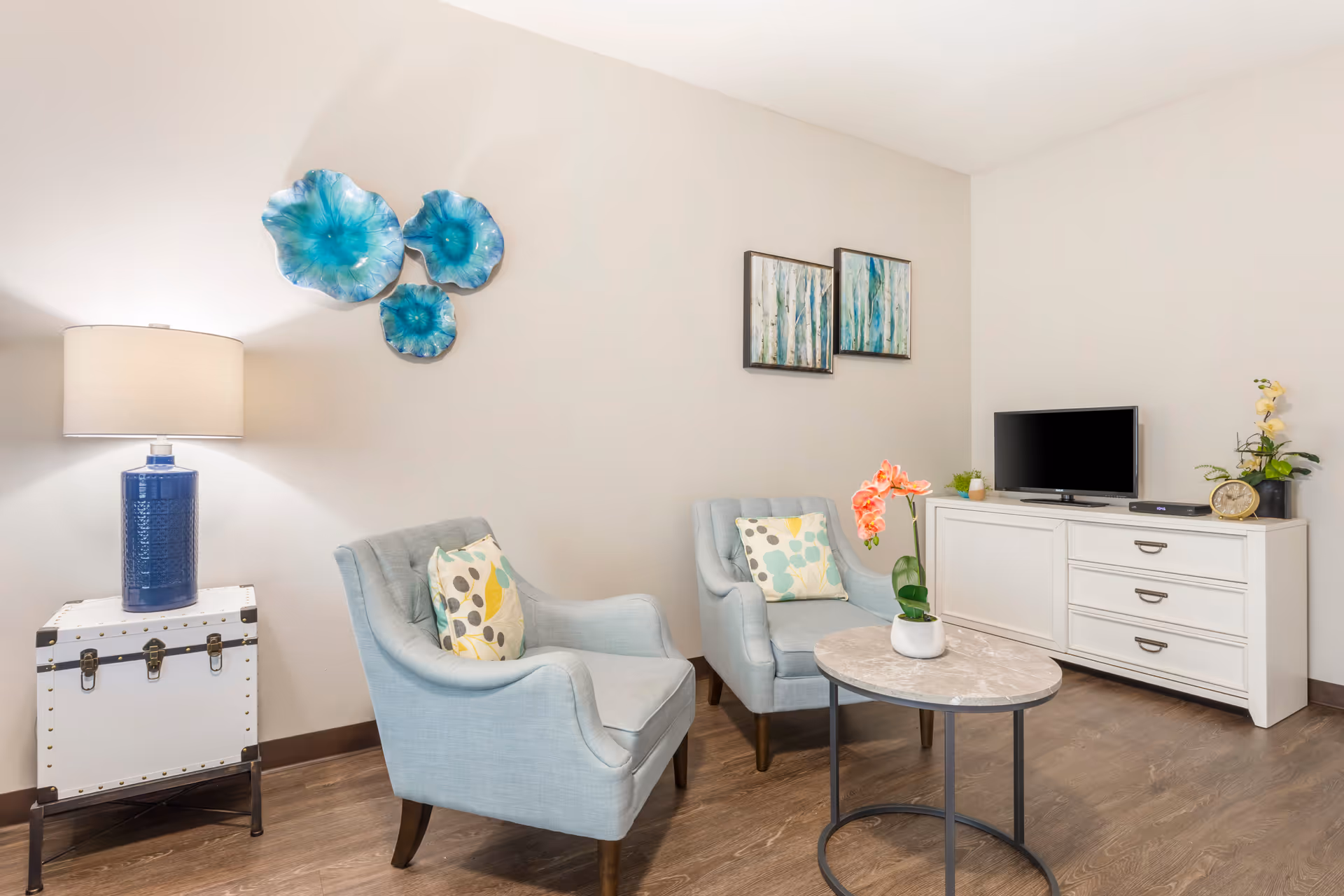 A cozy living room area with two light blue armchairs, each with a patterned cushion. Between the chairs is a round marble-top coffee table with a pink orchid plant. To the left is a white side table with a blue lamp. On the wall above the chairs are three blue decorative wall pieces and two framed paintings with blue and green tones. On the right side is a white TV stand with a flat-screen TV, a clock, and some small plants. The room has light-colored walls and wood flooring.