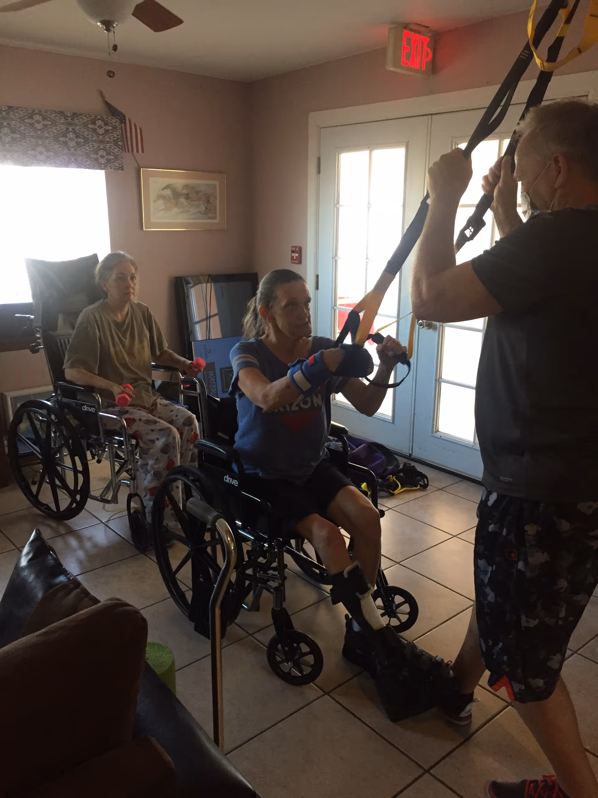 Two elderly women in wheelchairs participating in a physical therapy session indoors. One woman is using resistance bands held by a therapist, while the other woman holds small weights. The room has tiled floors, a ceiling fan, a framed picture on the wall, and a glass door with an exit sign above it.