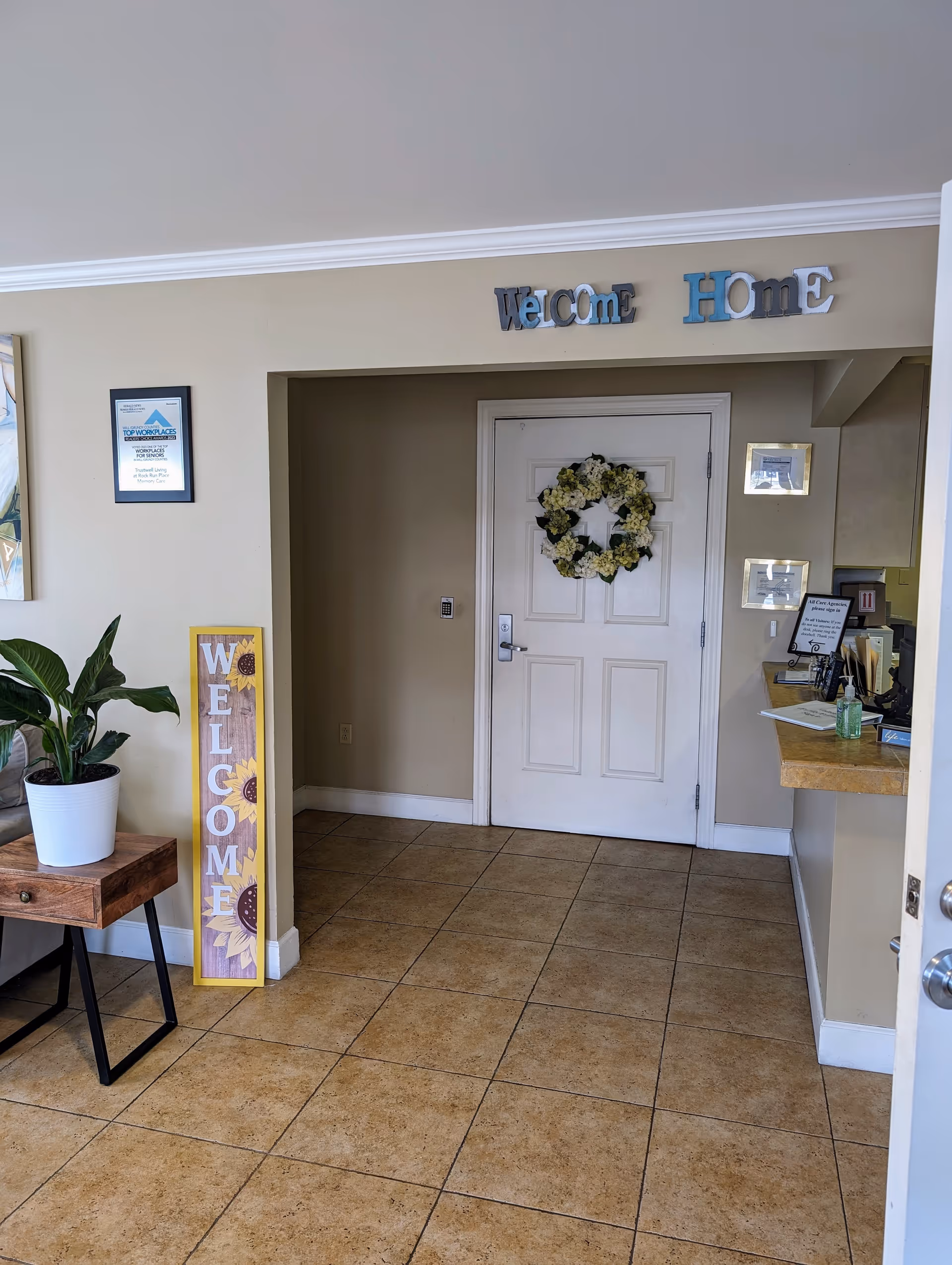 Small reception/entry area with a white door wreath, 'Welcome Home' letters above, a tall 'WELCOME' sign, and a potted plant on a side table.
