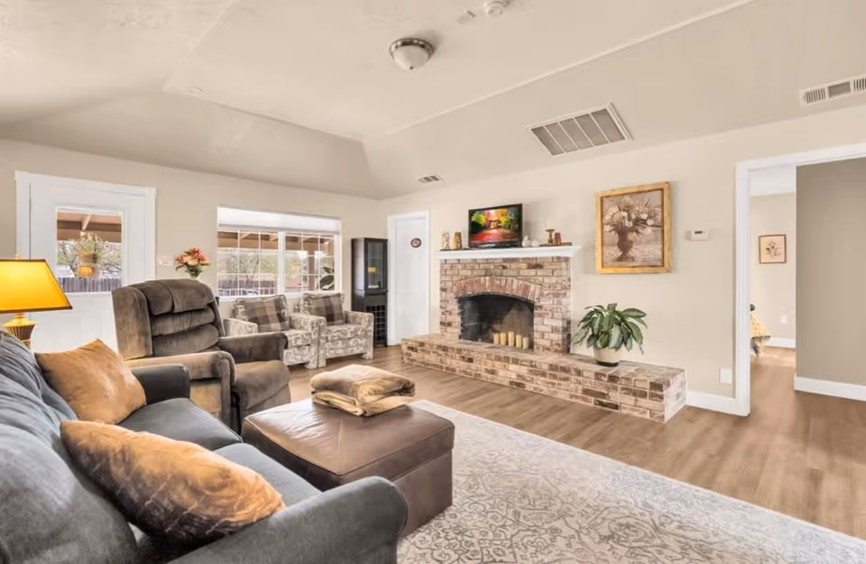 A cozy living room with a dark gray sofa, brown recliner chair, two patterned armchairs, and a brown ottoman. There is a brick fireplace with candles on the hearth and decorative items on the mantel, including a framed painting and a small TV. A potted plant sits on the hearth. The room has light-colored walls, wood flooring, a patterned area rug, and a window and door leading to an outdoor area.