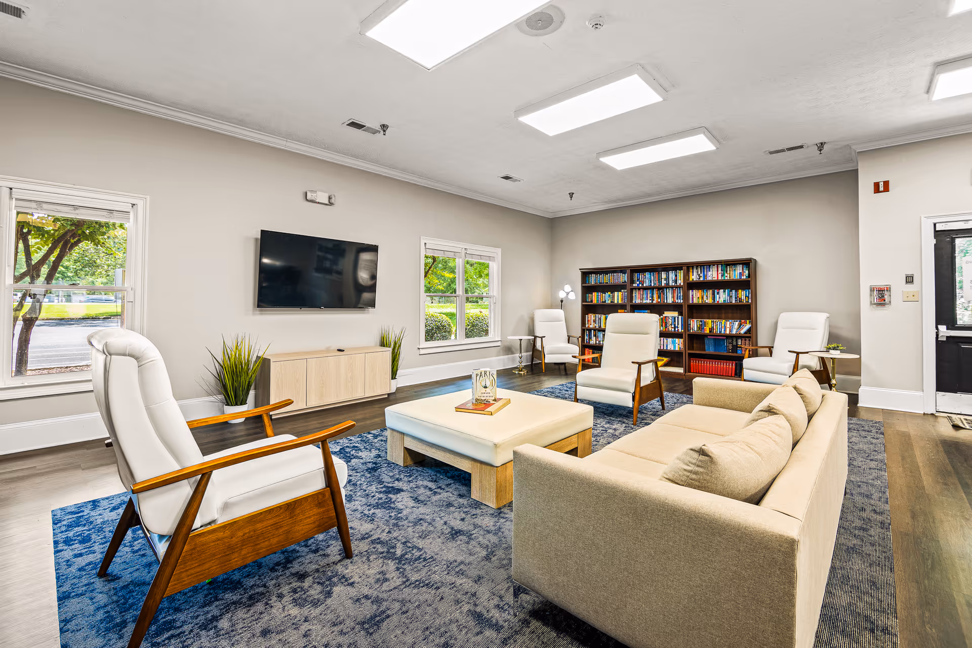 A bright and spacious living room in Avery Village Assisted Living and Memory Care featuring a beige sofa, four white cushioned armchairs with wooden frames, a large square ottoman with books on top, a blue area rug, a wall-mounted flat-screen TV, two windows showing greenery outside, and a bookshelf filled with books against the far wall.