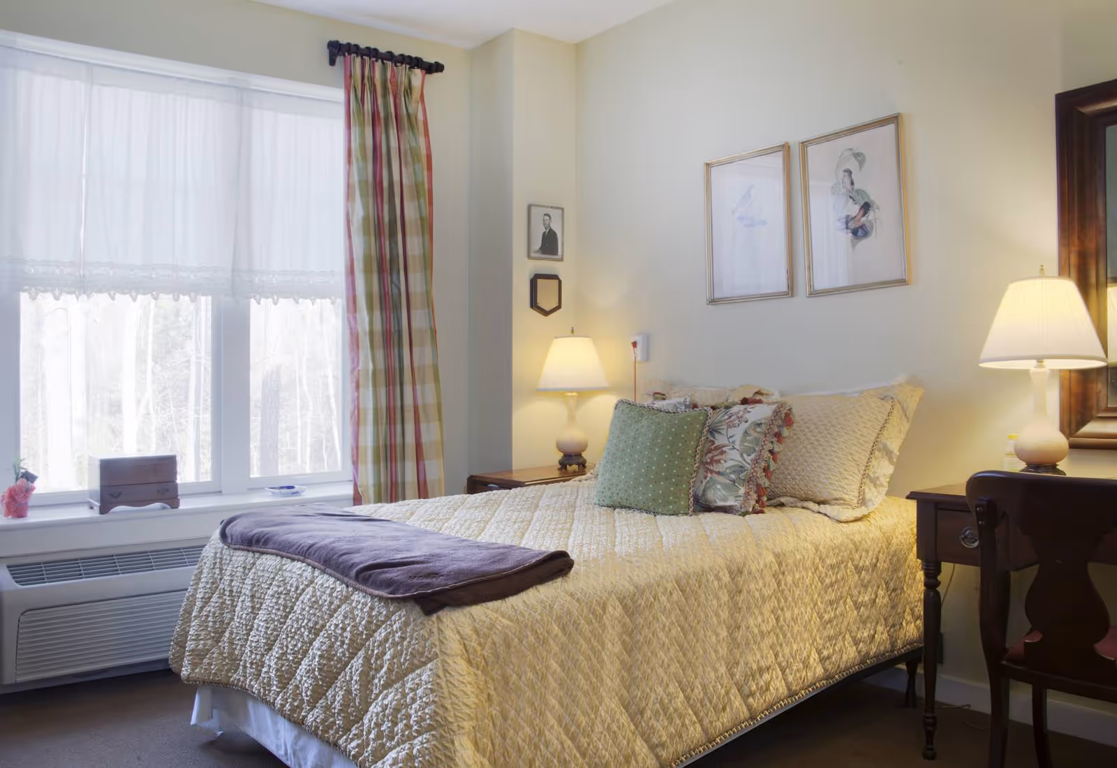 A cozy bedroom with a neatly made bed featuring a quilted beige bedspread and decorative pillows. There are two bedside tables, each with a lamp, and framed artwork on the wall above the bed. A window with sheer white curtains and plaid drapes allows natural light to fill the room.