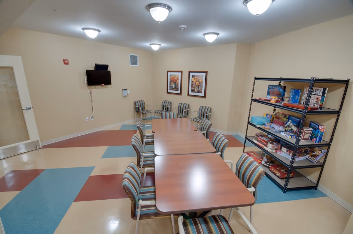 A senior living facility common room with three wooden tables arranged in a row, surrounded by striped chairs. On the right side, there is a black metal shelving unit filled with board games and puzzles. The walls are beige with two framed floral pictures. A flat-screen TV is mounted on the wall to the left, and the floor has a colorful geometric pattern with blue, beige, and red tiles. The room is lit by multiple ceiling lights.