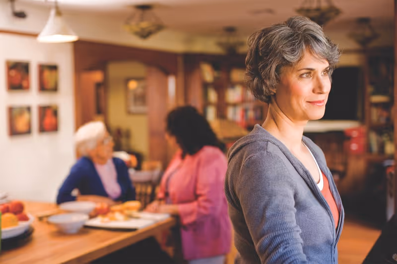 A middle-aged woman with short gray hair wearing a gray cardigan stands in the foreground of a cozy room. In the background, two elderly women are seated at a wooden table with bowls and fruit, engaged in conversation. The room has warm lighting, wooden furniture, and framed pictures on the wall.