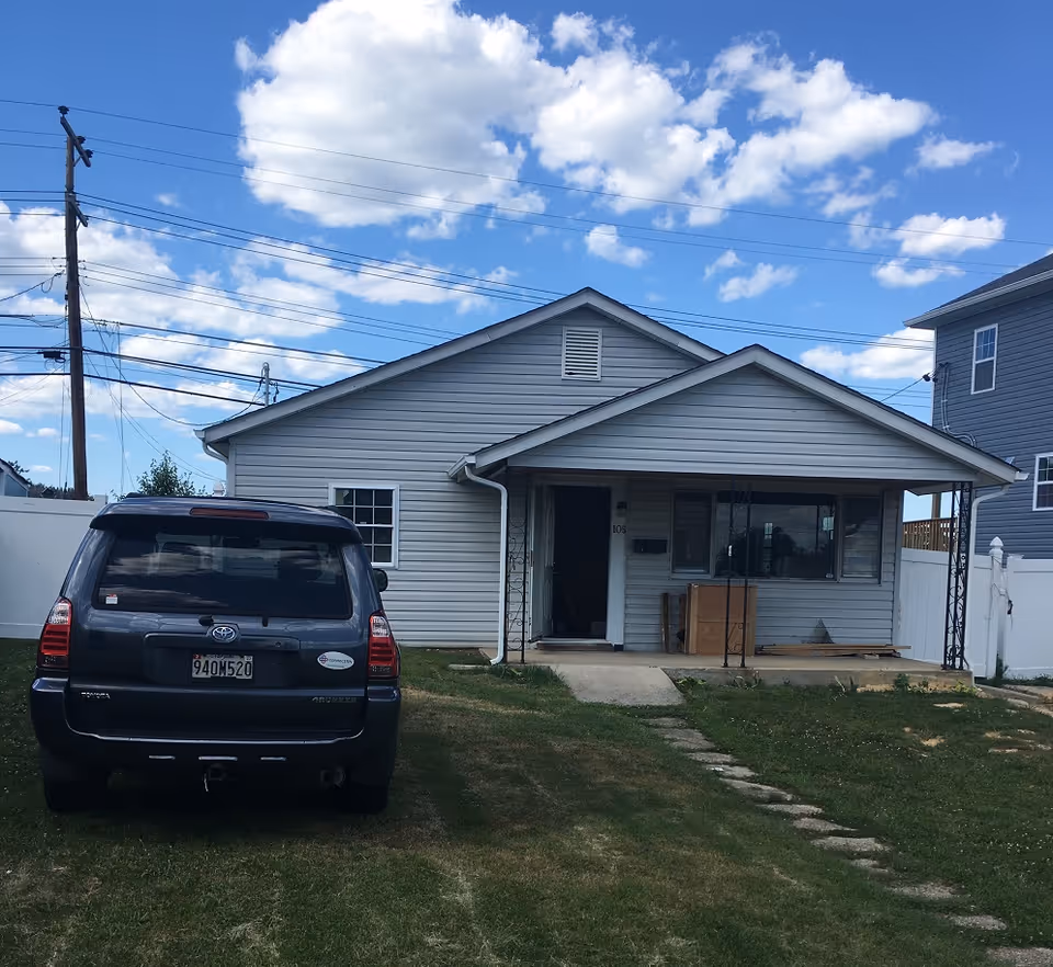Single-story house with gray siding and a covered front porch. A black SUV is parked on the grass driveway in front of the house. The sky is blue with scattered white clouds.