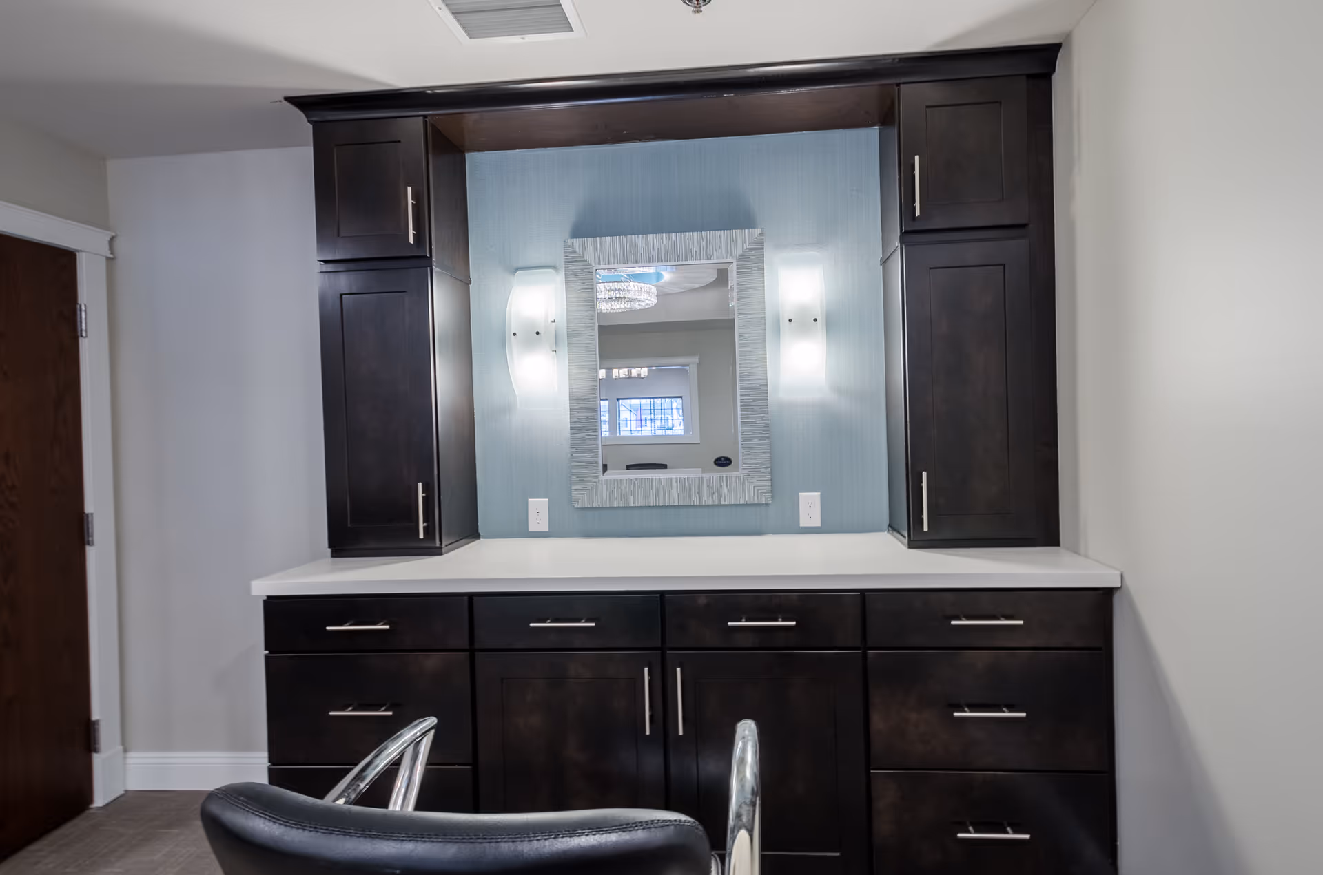 A vanity area with dark wood cabinets and drawers, a white countertop, a rectangular mirror with a textured frame, and two wall-mounted light fixtures on either side of the mirror. A black chair with chrome armrests is positioned in front of the vanity. The walls are light-colored, and there is a wooden door to the left.