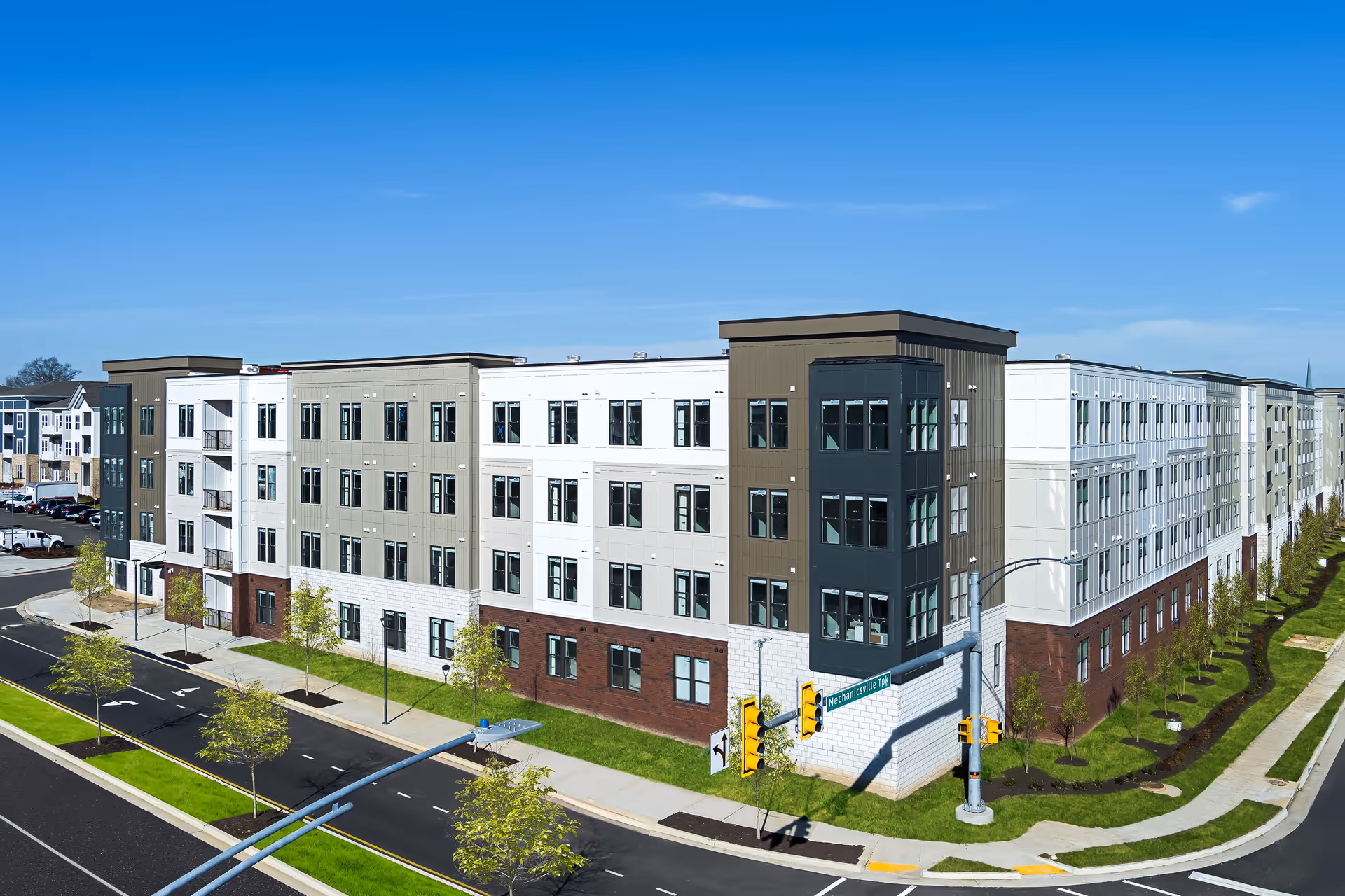Exterior view of a modern multi-story senior living facility building with a mix of white, gray, and brown panels, large windows, and a landscaped sidewalk with young trees along the street. A traffic light and street sign for Mechanicsville Tpk are visible at the corner.