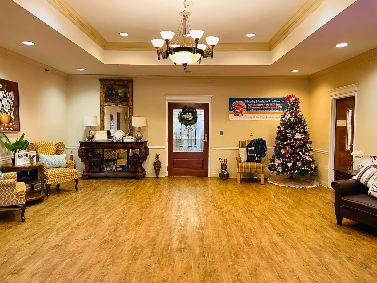 A warmly lit common area with wooden flooring, beige walls, and a tray ceiling with a chandelier. The room features several upholstered chairs, a wooden console table with lamps and a mirror, a decorated Christmas tree, and a door with a holiday wreath. There is a banner on the wall indicating the facility's name and an award received in 2019.