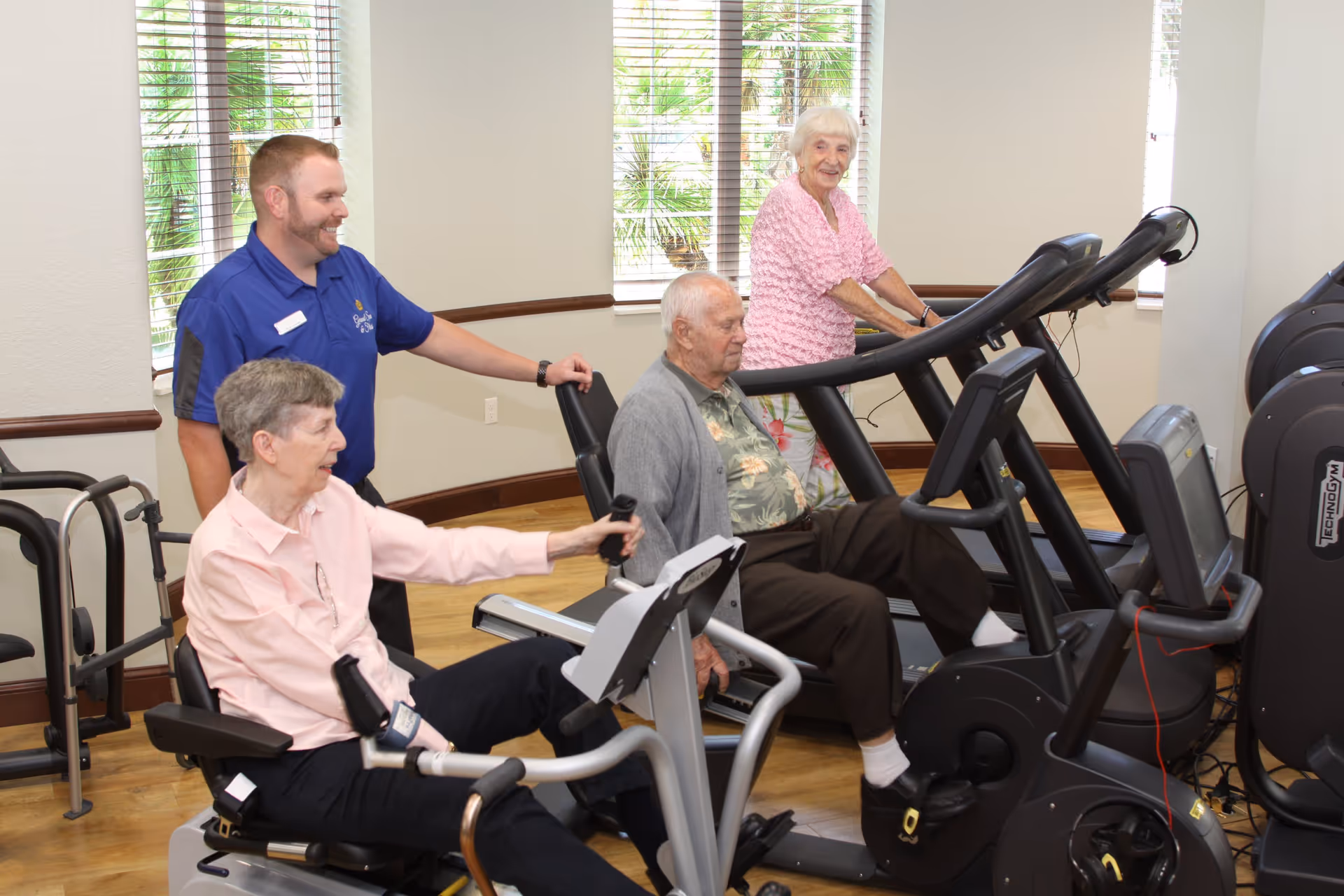 Three elderly individuals exercising on cardio machines in a fitness room with large windows and a staff member in a blue shirt assisting them.