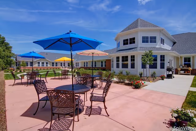 Outdoor patio with metal tables, chairs and colorful umbrellas beside a light-colored multi-windowed senior living building under a blue sky.