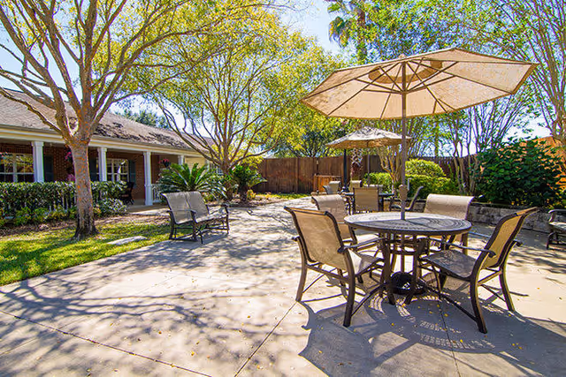 Outdoor patio area with round tables and chairs under large beige umbrellas, surrounded by trees and greenery. A building with a covered porch and windows is visible in the background.