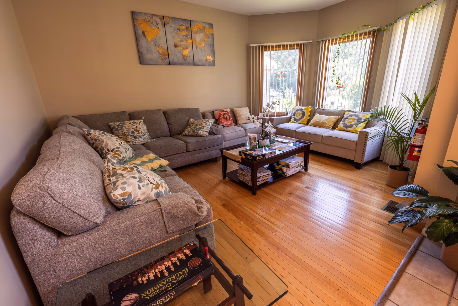 A cozy living room with two gray sofas adorned with floral and patterned cushions, a wooden coffee table with board games and magazines underneath, large windows with vertical blinds letting in natural light, a potted plant near the window, and a three-panel world map artwork on the wall.