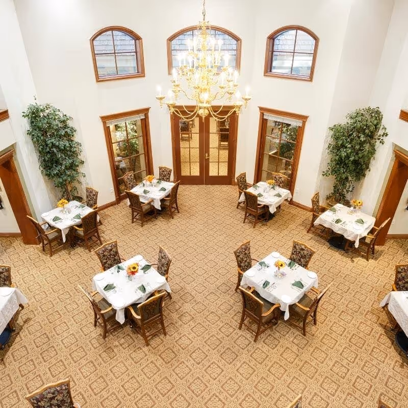 Elegant dining room with multiple small tables set for meals, patterned carpet, tall windows and a central chandelier.