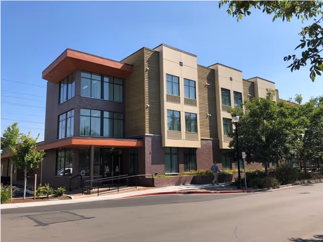 Exterior view of a modern three-story building with large windows and a mix of brick and siding materials. There is a sidewalk with a person walking, trees, and a clear blue sky.