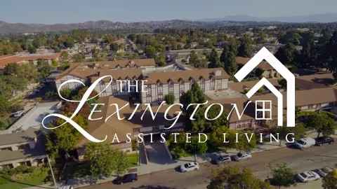 Aerial view of Lexington Assisted Living facility showing multiple buildings surrounded by trees and parked cars, with mountains visible in the background under a clear sky.