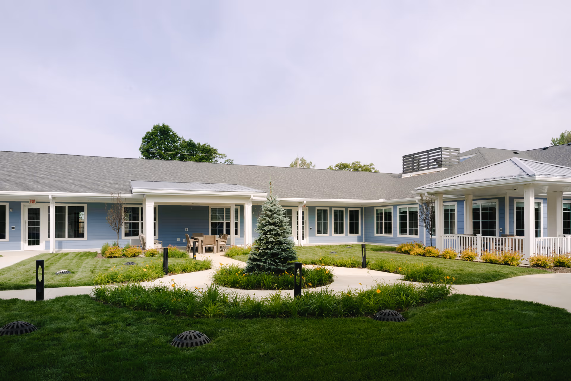 Outdoor courtyard area of a senior living facility with a well-maintained lawn, a small evergreen tree in the center, paved walkways, and a blue building with white trim surrounding the courtyard. There is a covered patio with outdoor seating and tables.