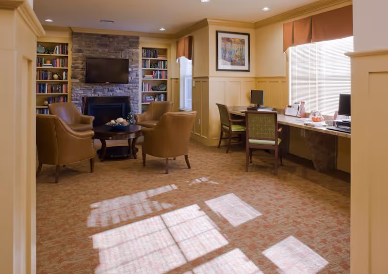 A cozy common area with four brown leather armchairs arranged around a wooden coffee table in front of a stone fireplace with a mounted flat-screen TV. Built-in bookshelves flank the fireplace, filled with books and decorative items. To the right, there is a long desk with two chairs and computers, positioned under windows with blinds and valances. The room has warm yellow walls with wainscoting and carpeted flooring.