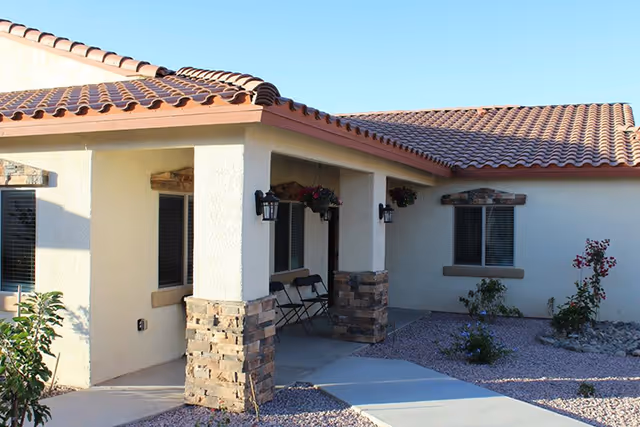 Exterior view of a single-story building with a tiled roof and beige stucco walls. The entrance area features stone pillars, hanging flower baskets, and a few folding chairs. There are windows with blinds and a landscaped area with small plants and gravel.