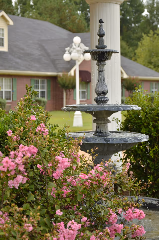 A three-tiered black water fountain surrounded by pink flowering bushes in the garden area of a senior care facility. In the background, there is a brick building with green window shutters and a white column with a decorative street lamp.