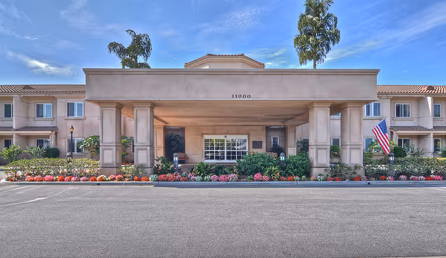 Front entrance of a two-story senior living facility with a covered porte-cochère, landscaped flowerbeds, and an American flag.