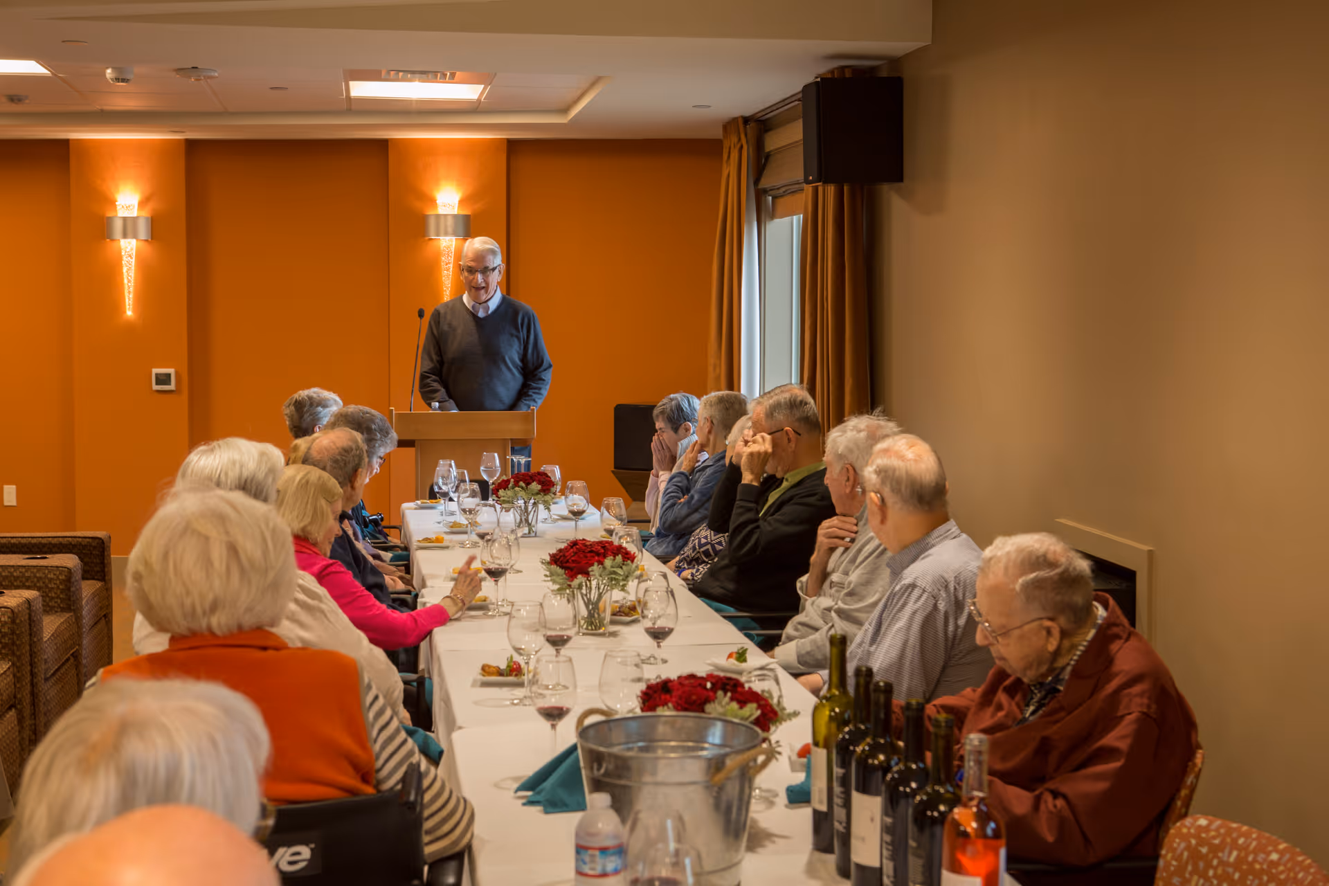 A group of elderly people seated around a long dining table with wine glasses and floral centerpieces, listening to a man standing at a podium speaking in a warmly lit room with orange walls and wall sconces.
