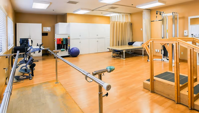 Physical therapy room with parallel bars, exercise equipment, a therapy table, and a small practice staircase.