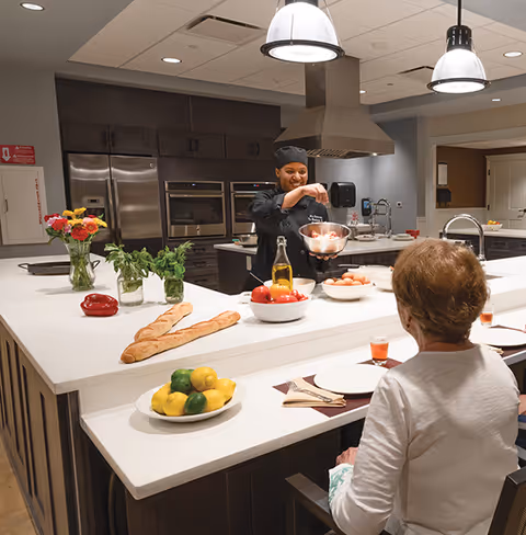 A chef in a black uniform is preparing food behind a large kitchen island in a modern kitchen. In the foreground, an elderly woman is seated at the counter with a plate and glass in front of her. The kitchen features stainless steel appliances, including ovens and a refrigerator, and is decorated with fresh flowers, herbs, bread, lemons, and tomatoes on the counter.