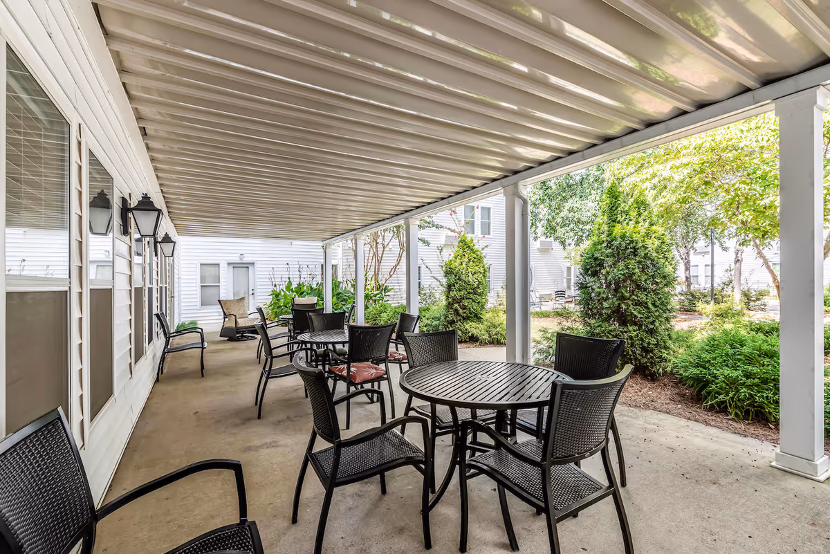 Covered outdoor patio area with several black metal tables and chairs arranged for seating. The patio is adjacent to a white building with windows and outdoor wall lanterns. Green shrubs and trees are visible beyond the patio area.