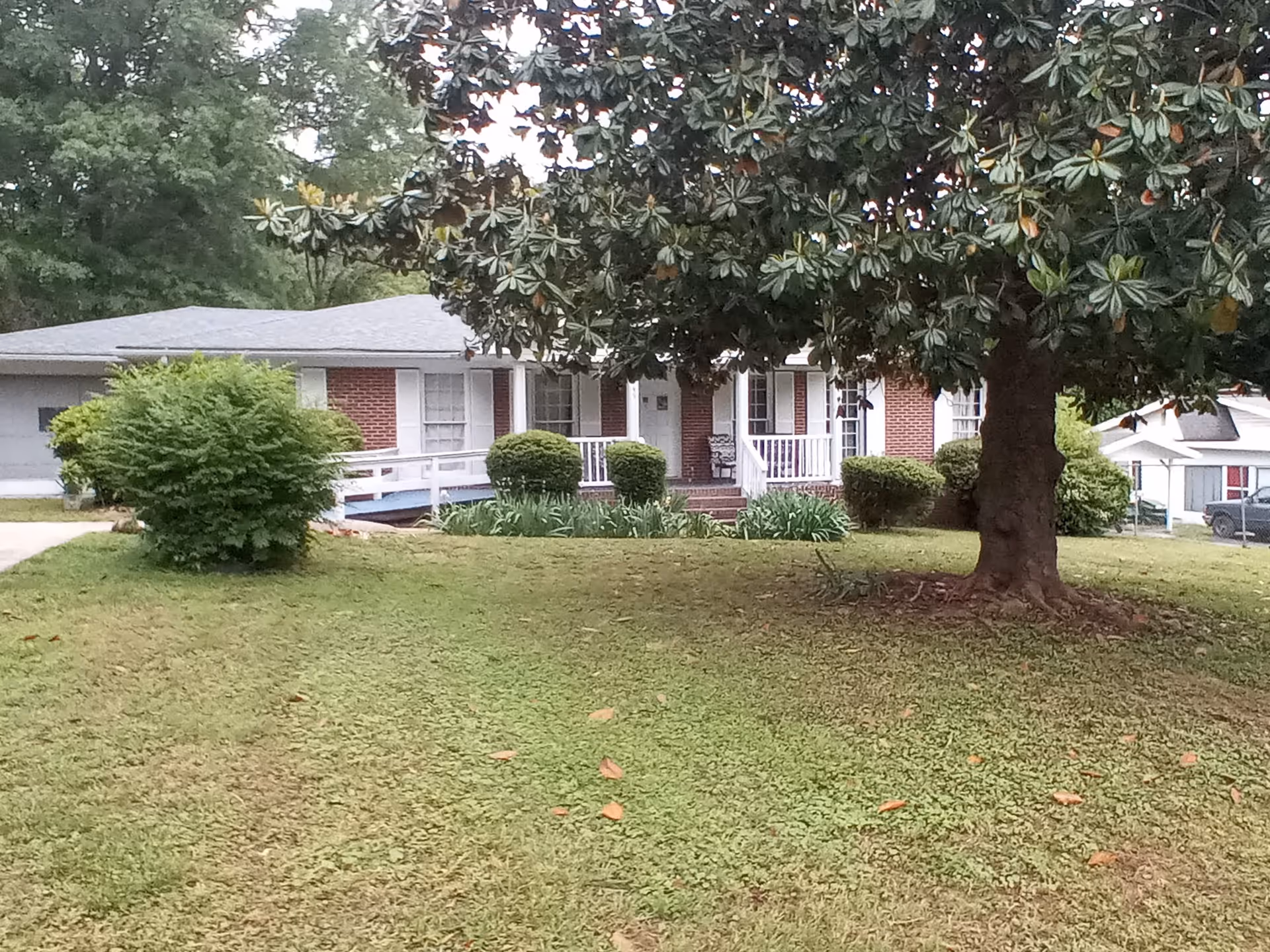 Front exterior view of a single-story brick house with white trim, a porch with white railings, a wheelchair ramp, and a large tree in the front yard with green grass and bushes.