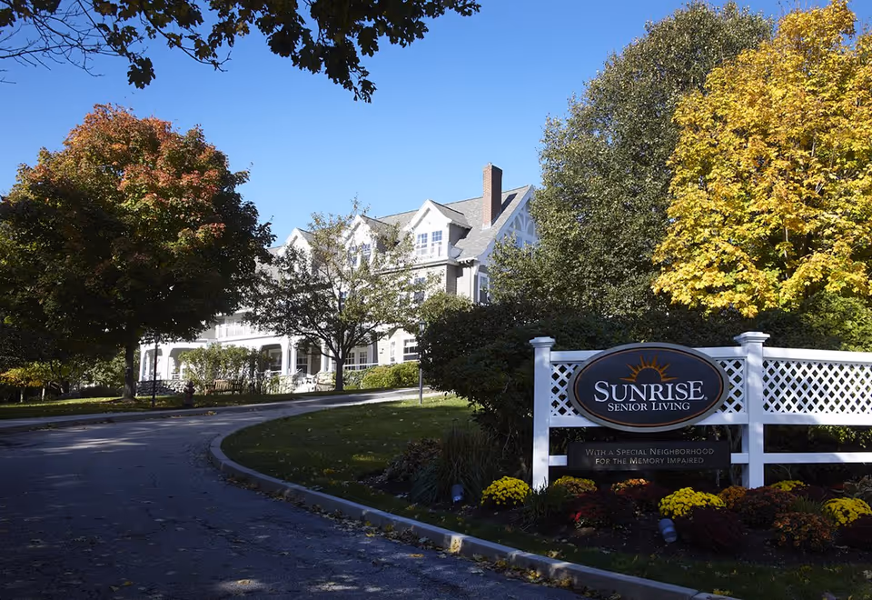 Entrance driveway and front of a white senior living building with a 'Sunrise Senior Living' sign and autumn trees.