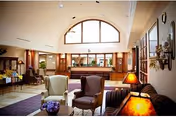 Lobby-style common room with upholstered chairs and sofas, table lamps, floral arrangements, and a reception desk beneath a large arched window.