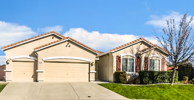 Exterior view of a single-story residential building with beige walls, two garage doors, a driveway, green lawn, bushes, and a tree under a blue sky with some clouds.
