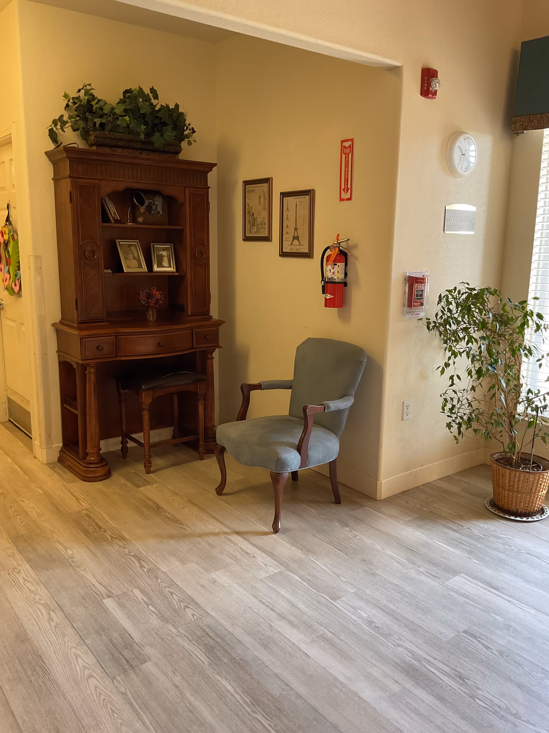 A cozy corner in an assisted living facility featuring a wooden hutch with framed photos and decorative items, a cushioned armchair with wooden arms, a potted plant near a window with blinds, and safety equipment including a fire extinguisher and fire alarm on the wall.