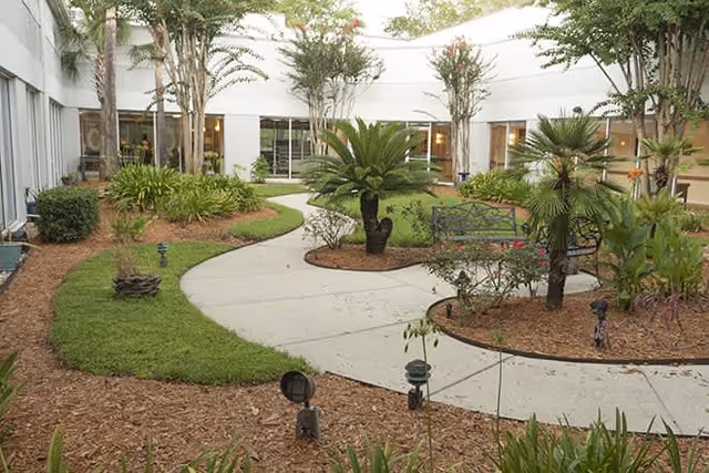 A landscaped outdoor courtyard with a curved concrete pathway surrounded by green grass, various plants, small trees, and mulch beds. There are benches along the pathway and large windows of the building visible in the background.