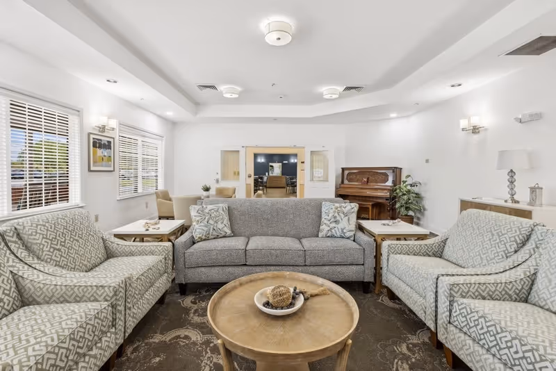 A bright and spacious living room area with a gray sofa and two patterned armchairs arranged around a round wooden coffee table. The room features large windows with white blinds, a piano in the corner, and a doorway leading to another room with a dining table and chairs. The walls are white, and the ceiling has recessed lighting and modern light fixtures.