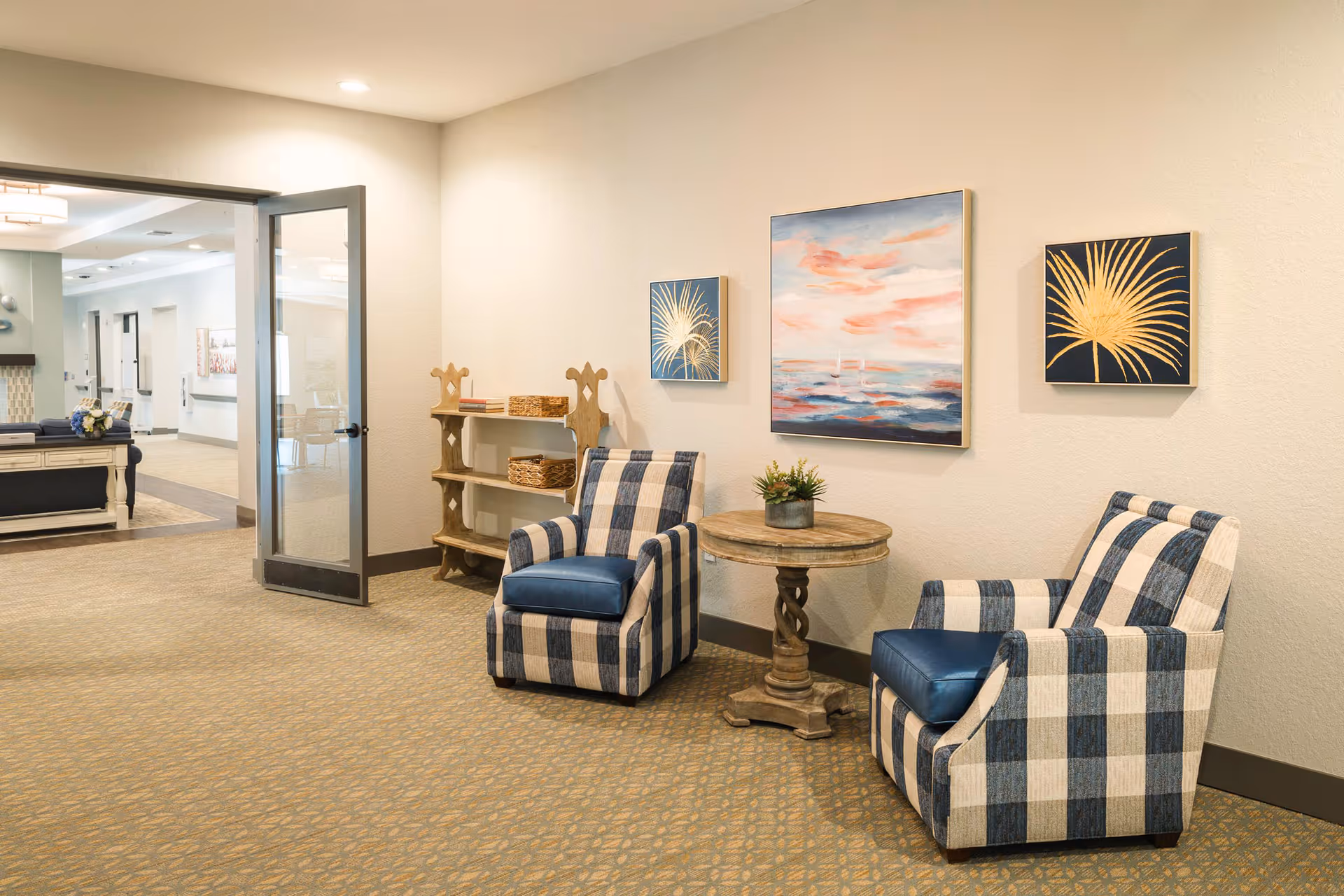 A cozy seating area in a senior living facility with two blue and white checkered armchairs positioned around a small round wooden table with a potted plant. The wall behind features three framed artworks, including a large painting of a sunset over water and two smaller pieces with gold palm leaf designs. To the left, an open glass door leads to a hallway with additional seating and decor.