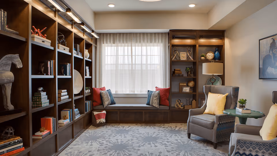 A cozy reading room with built-in dark wood bookshelves filled with books and decorative items. A window seat with cushions is centered under a large window with sheer curtains. Two gray armchairs with yellow pillows flank a small round green table with a plant and books. The room has a patterned carpet and soft lighting.