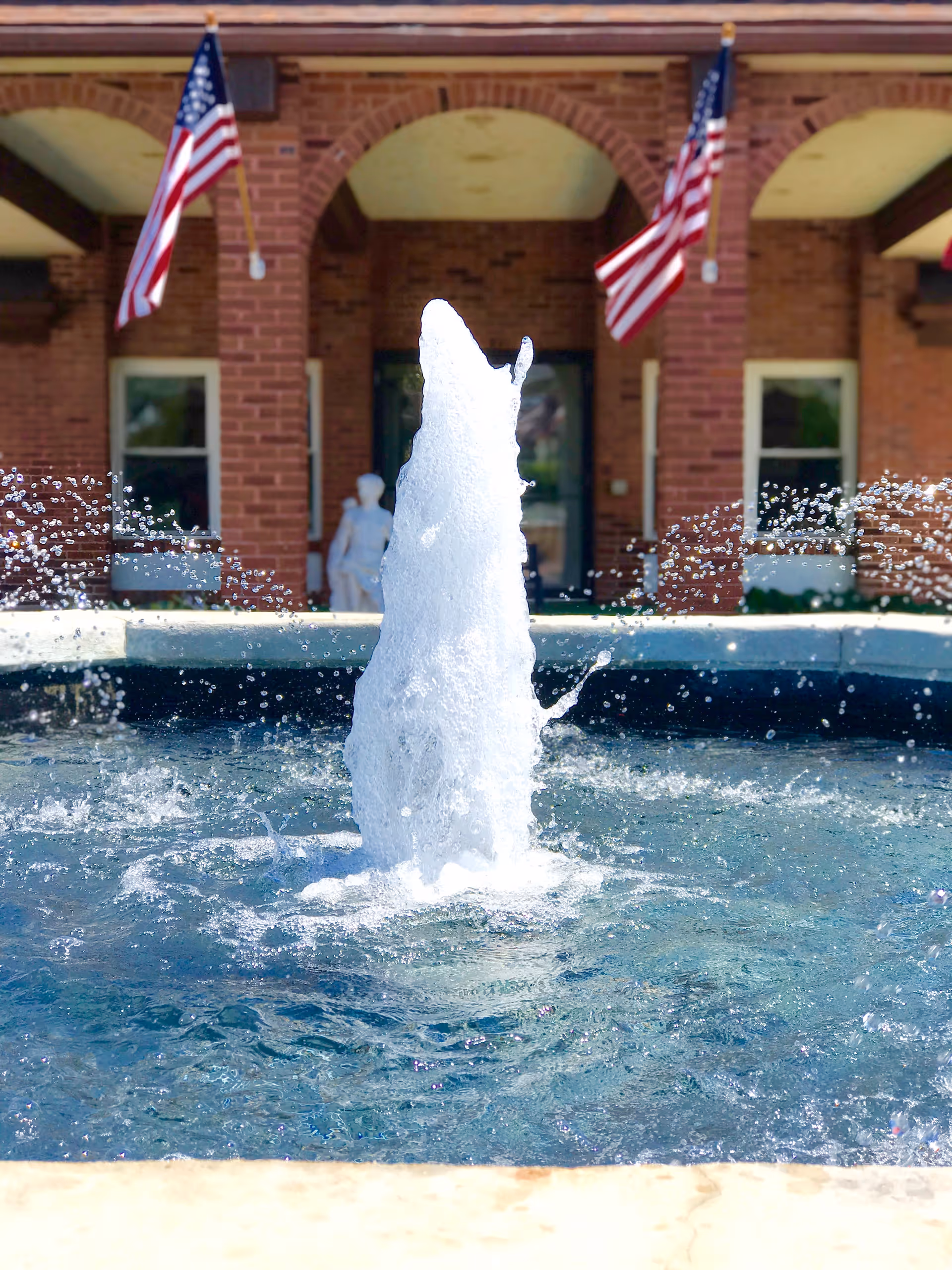 A water fountain with water spraying upwards in front of a brick building entrance with two American flags hanging on either side of the doorway.
