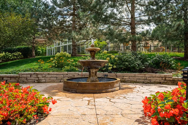 Outdoor garden area with a two-tiered stone water fountain in the center, surrounded by a stone-paved circular patio. There are vibrant red flowers in the foreground on both sides, green bushes, and tall pine trees in the background with a greenhouse structure partially visible behind the trees.