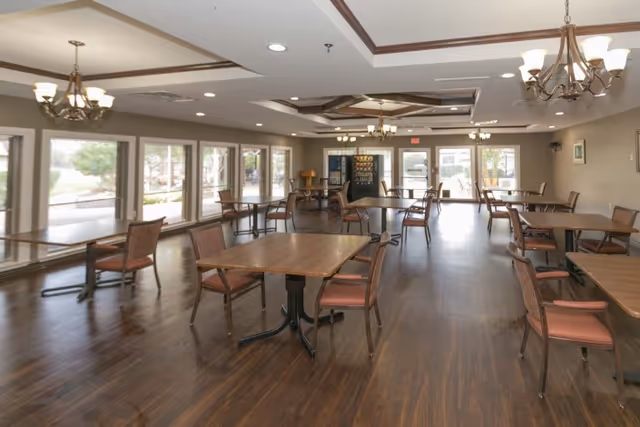 A spacious dining room with multiple wooden tables and chairs arranged neatly. Large windows line one side of the room, allowing natural light to fill the space. There are chandeliers hanging from the ceiling, and vending machines are visible at the far end near the exit doors.